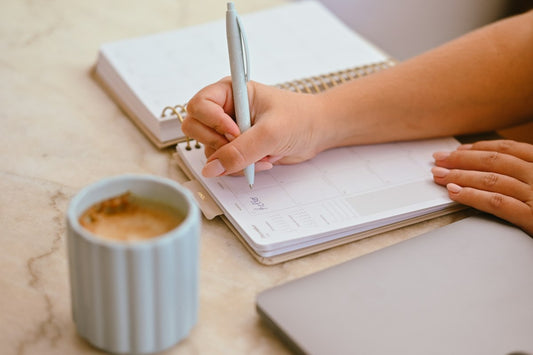 A person writing in a planner with a light blue pen next to a coffee mug